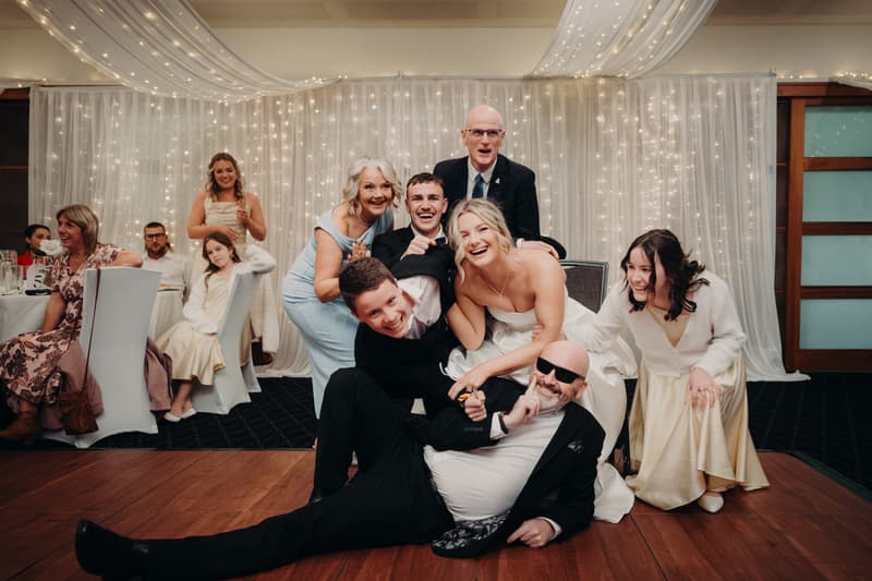 Bride Libby and groom Kyle pose with family members and guests in front of a white curtain with string lights at The Tides — The Pandanus Room during the reception.