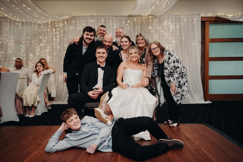 Bride Libby in a white strapless gown and groom Kyle in a black tuxedo sit together in The Pandanus Room at The Tides, surrounded by family and guests posing for a group photo at the reception. A young boy lies on the floor in front of them, and other guests are seated at tables in the background.