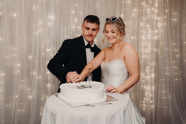 Bride Libby and groom Kyle cut their wedding cake together at The Tides — The Pandanus Room, standing behind a round table with a white tablecloth and a white cake, with a backdrop of sheer curtains and string lights.
