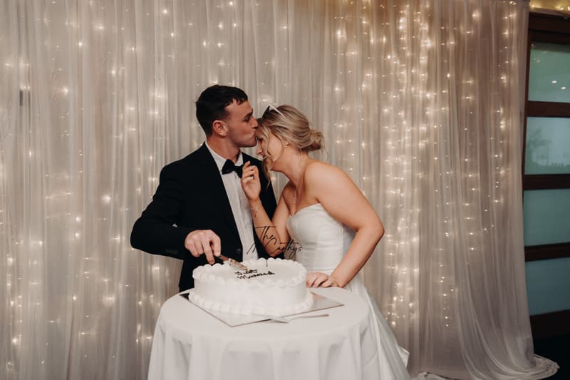 Bride Libby and groom Kyle stand at a round table cutting their wedding cake at The Tides — The Pandanus Room, with a backdrop of white curtains and string lights.