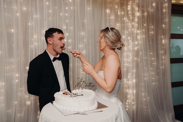 Bride Libby feeds a piece of wedding cake to groom Kyle at their reception in The Tides — The Pandanus Room, with a backdrop of string lights and sheer curtains.