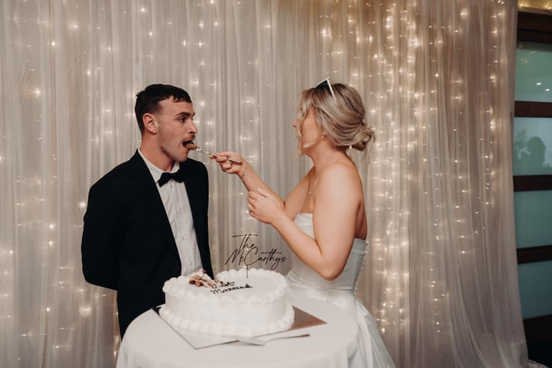 Bride Libby feeds a piece of wedding cake to groom Kyle at their reception in The Tides — The Pandanus Room, with a backdrop of string lights and sheer curtains.
