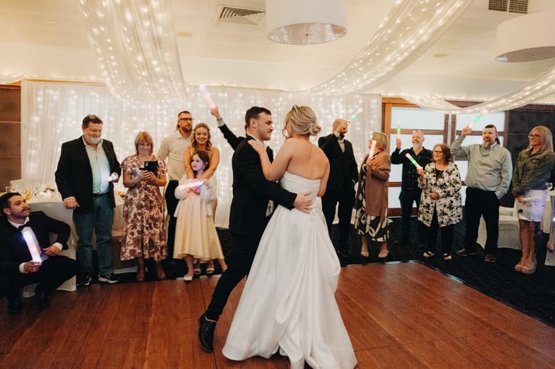 Bride Libby and groom Kyle share a dance on the wooden dance floor at The Tides — The Pandanus Room, surrounded by guests holding glowing light sticks and watching.