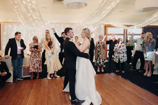 Bride Libby and groom Kyle share a dance on the wooden dance floor at The Tides — The Pandanus Room, surrounded by guests holding light sticks and watching.