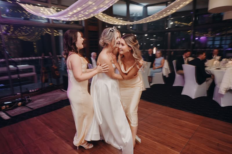 The bride Libby embraces two bridesmaids on the dance floor at The Tides — The Pandanus Room during the wedding reception.