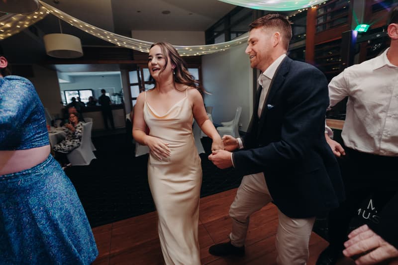 The bride Libby in a cream satin dress and the groom Kyle in a dark blazer and light pants dance together at the reception in The Tides — The Pandanus Room, with guests seated at tables in the background.