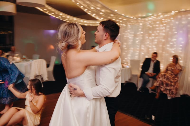 Bride Libby and groom Kyle share a dance at the reception in The Pandanus Room at The Tides, with guests seated and watching in the background under string lights.