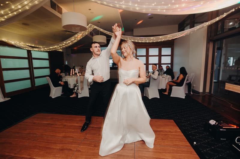 The bride Libby and groom Kyle dance on the wooden dance floor at The Tides — The Pandanus Room during the wedding reception, with guests seated at tables in the background.