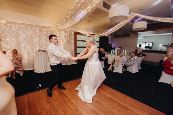 The bride Libby and groom Kyle hold hands and dance on the wooden dance floor at The Tides — The Pandanus Room, while guests sit at tables watching.
