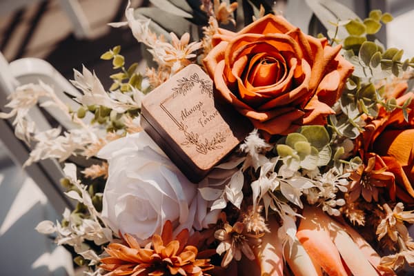 Close-up of a wedding bouquet featuring orange and white roses, assorted dried flowers, and a wooden ring box engraved with 'Always & Forever'. Valentine's Day wedding photography by She Said Yes.