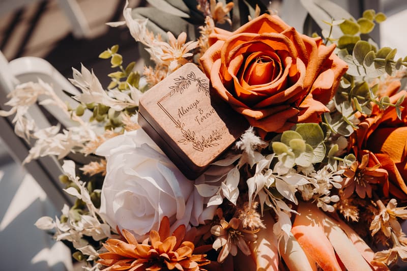 Close-up of a wedding bouquet featuring orange and white roses, assorted dried flowers, and a wooden ring box engraved with 'Always & Forever'. Brisbane wedding photographers by She Said Yes.