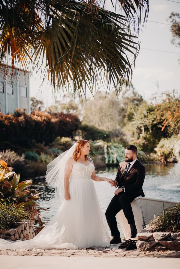 The bride Lilas in a white wedding gown and veil stands holding hands with the groom Kaine, who is seated on the edge of a small boat by a pond at Eatons Hill Hotel, surrounded by greenery and palm fronds overhead.