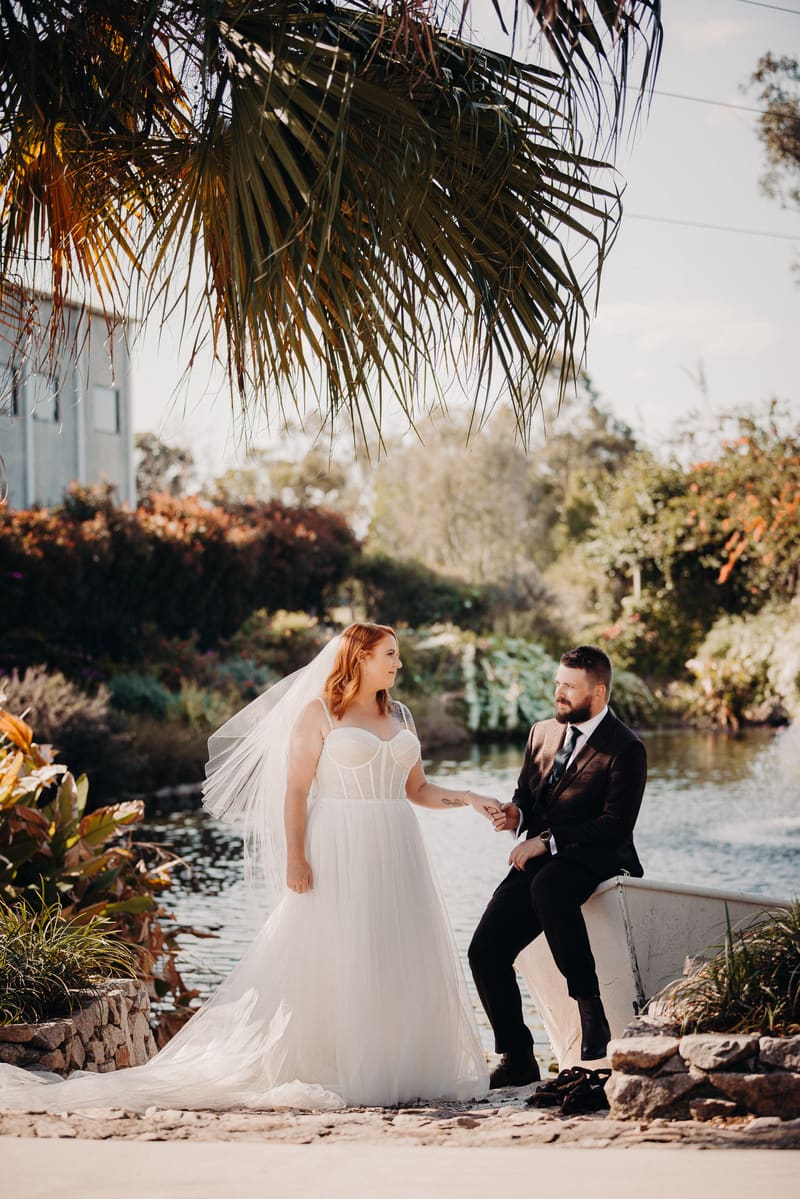The bride Lilas in a white wedding gown and veil stands holding hands with the groom Kaine, who is seated on the edge of a small boat by a pond, surrounded by greenery and palm fronds overhead. Winter wedding venues Brisbane indoor by She Said Yes.