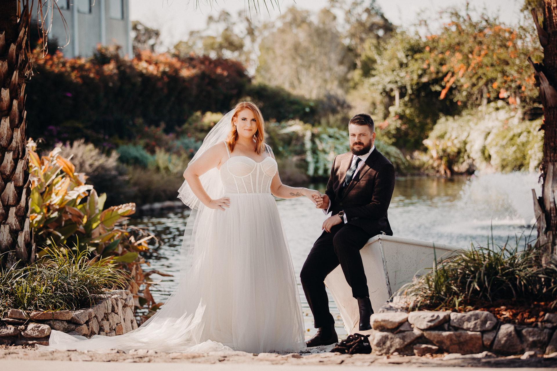 The bride Lilas stands beside the groom Kaine who is seated on the edge of a small boat by a pond at Eatons Hill Hotel, holding hands with greenery and a water fountain in the background.