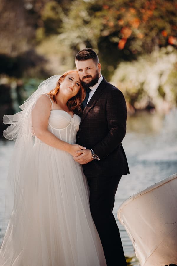 The bride Lilas in a white wedding gown and veil leans her head on the groom Kaine, who is wearing a dark suit and tie, as they stand close together near a body of water at Eatons Hill Hotel.
