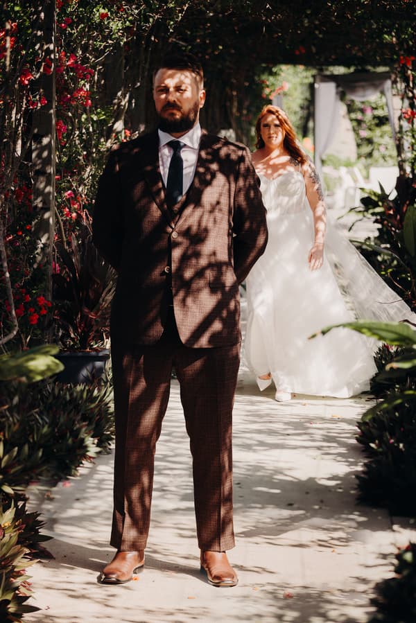 The groom Kaine stands with his eyes closed under dappled sunlight on a garden path at Eatons Hill Hotel, while the bride Lilas walks towards him from behind in her wedding gown.