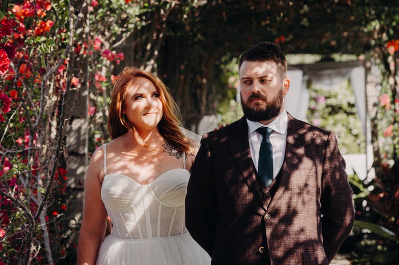 The bride Lilas in a white strapless wedding dress and the groom Kaine in a dark suit with a tie stand together under a floral archway at Eatons Hill Hotel during their couple portraits session.