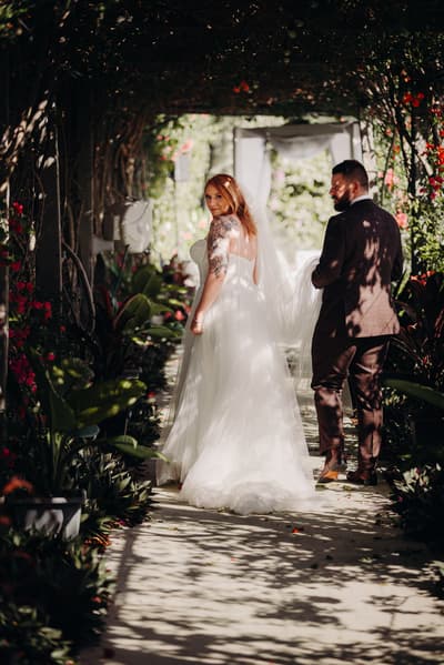 The bride Lilas in a white wedding gown and the groom Kaine in a dark suit walk together under a shaded archway with greenery and flowers. Lilas looks back towards the camera. Winter wedding Brisbane dress code by She Said Yes.