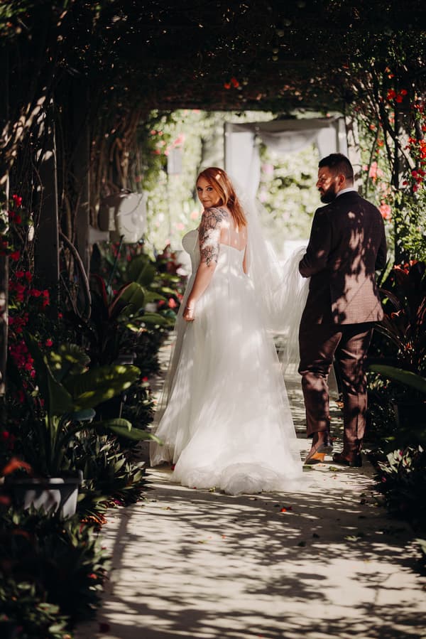The bride Lilas in a white wedding gown and the groom Kaine in a dark suit walk together under a shaded archway with greenery and flowers at Eatons Hill Hotel. Lilas looks back towards the camera.