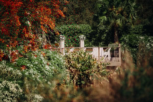 Empty ceremony stage decorated with floral arrangements on pillars and white chairs at Eatons Hill Hotel — Lakeside, surrounded by lush greenery and vibrant red and white flowers.