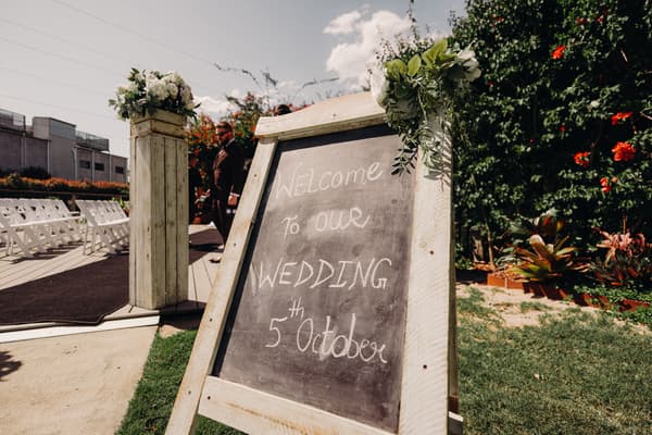 A chalkboard sign reading 'Welcome to our wedding 5th October' is displayed outdoors near white chairs arranged for a ceremony at Eatons Hill Hotel — Lakeside.