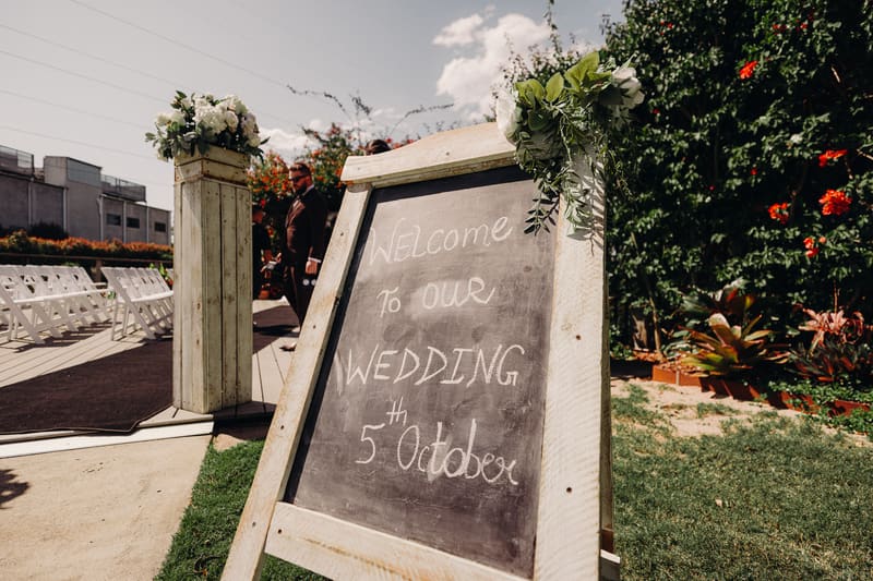 A chalkboard sign reading 'Welcome to our wedding 5th October' is displayed outdoors near white chairs arranged for a ceremony. Wedding fair season Brisbane by She Said Yes.