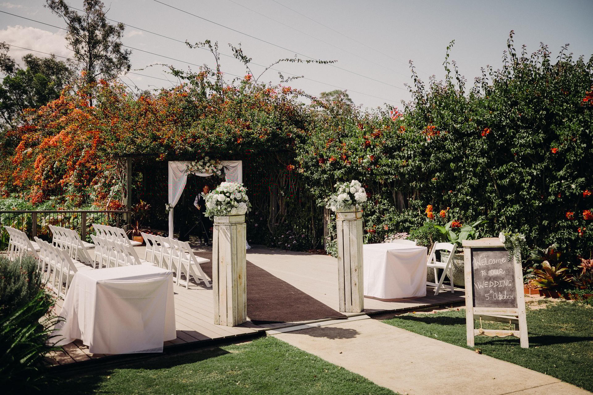 Outdoor wedding ceremony setup at Eatons Hill Hotel — Lakeside with white chairs arranged in rows, floral pillars, a decorated arbor, and a chalkboard sign welcoming guests.