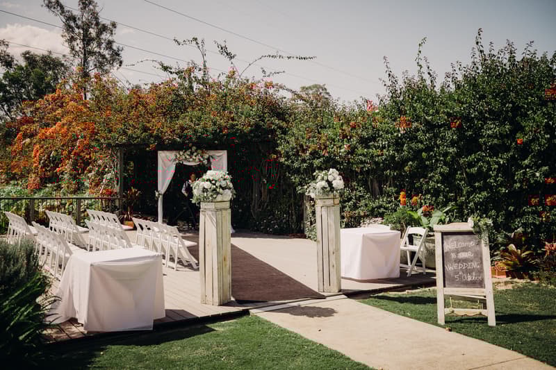 Outdoor wedding ceremony setup at Eatons Hill Hotel — Lakeside with white chairs arranged in rows, floral pillars, a decorated arbor, and a chalkboard sign welcoming guests.