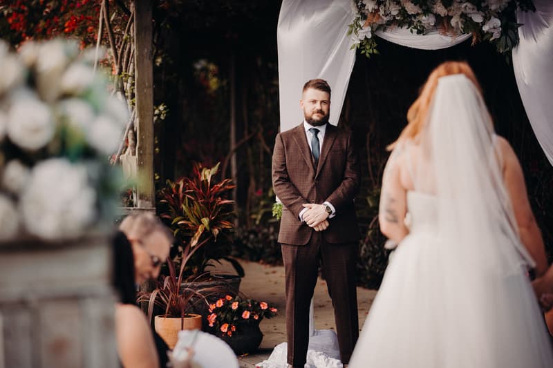 The groom Kaine stands under a floral arch at Eatons Hill Hotel — Lakeside, facing the bride Lilas who is walking towards him during the ceremony.