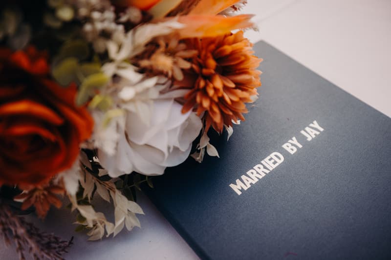 Close-up of a wedding bouquet with orange and white flowers resting on a navy blue book titled 'MARRIED BY JAY'.