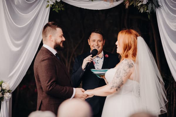 Bride Lilas and groom Kaine hold hands facing each other during their wedding ceremony at Eatons Hill Hotel — Lakeside, with the officiant speaking into a microphone between them under a floral and fabric arch.