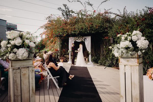 Bride Lilas and groom Kaine stand holding hands under a floral arch at the ceremony stage at Eatons Hill Hotel — Lakeside, with guests seated on either side of the aisle.