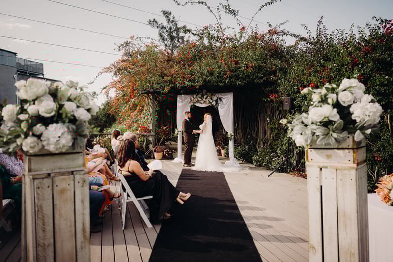 Bride Lilas and groom Kaine stand holding hands under a floral arch at the ceremony stage at Eatons Hill Hotel — Lakeside, with guests seated on either side of the aisle.