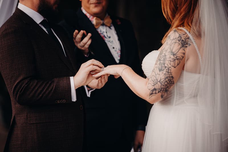 Kaine places a wedding ring on Lilas's finger during the ceremony at Eatons Hill Hotel — Lakeside, with the officiant standing behind them.