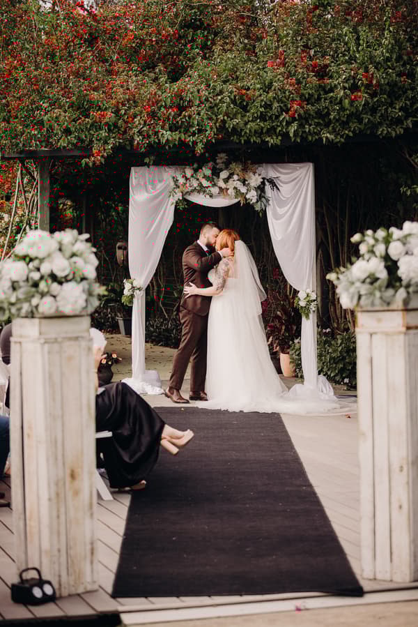 The bride Lilas and groom Kaine share a kiss under a floral arch draped with white fabric at the ceremony stage at Eatons Hill Hotel — Lakeside, with guests seated along the aisle.