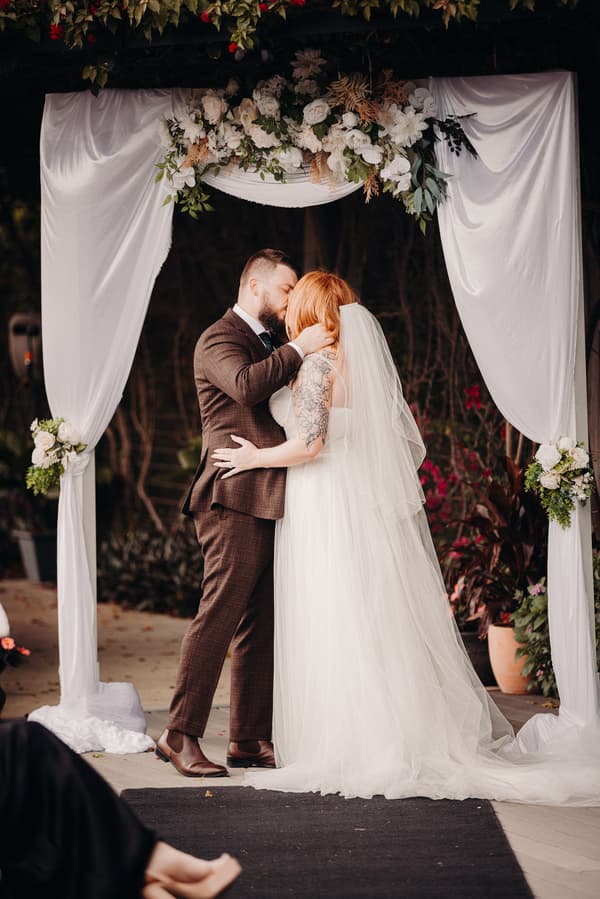 The bride Lilas and groom Kaine share a kiss under a floral and white fabric wedding arch at Eatons Hill Hotel — Lakeside during their ceremony.