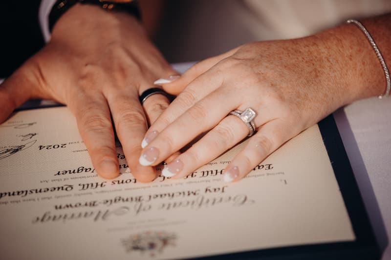 The bride and groom place their hands together on a wedding certificate at Eatons Hill Hotel — Lakeside, showing their wedding rings.