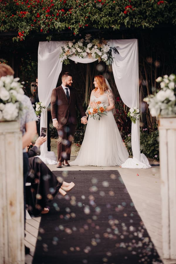 Bride Lilas and groom Kaine hold hands under a floral arch draped with white fabric at the ceremony stage at Eatons Hill Hotel — Lakeside, with guests seated along the aisle.