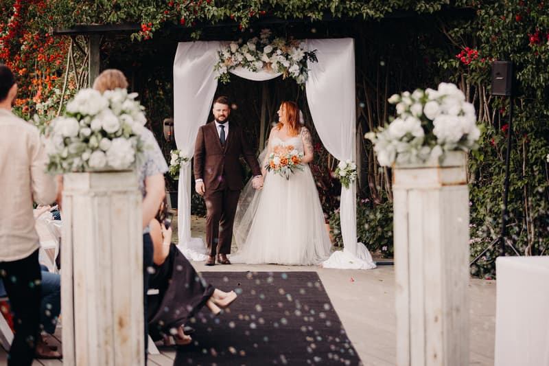 The bride Lilas and groom Kaine hold hands standing under a white floral arch at Eatons Hill Hotel — Lakeside during their wedding ceremony, with guests seated on either side of the aisle.