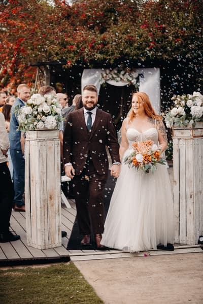 Bride Lilas and groom Kaine walk hand-in-hand down the aisle at Eatons Hill Hotel — Lakeside, surrounded by guests and bubbles, with floral arrangements on white wooden pillars.