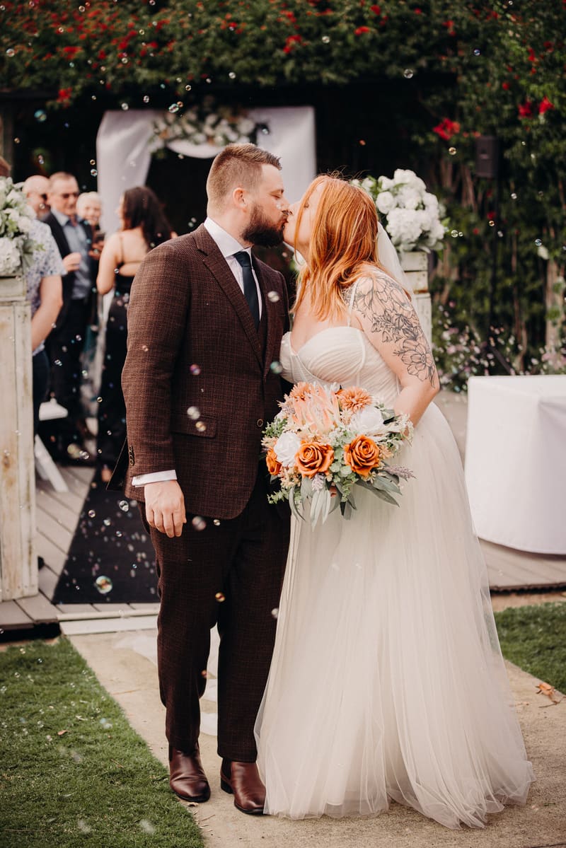 Bride Lilas and groom Kaine kiss at the ceremony stage, surrounded by guests and floral decorations, with bubbles floating in the air. Wedding date changed COVID by She Said Yes.