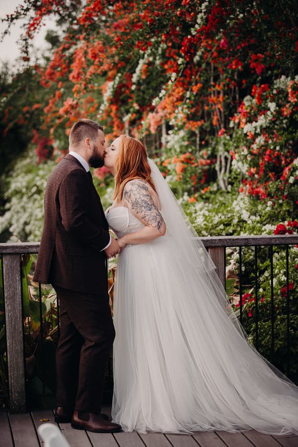 Bride Lilas and groom Kaine share a kiss while holding hands on a wooden deck at Eatons Hill Hotel, with a backdrop of vibrant red and white flowers.
