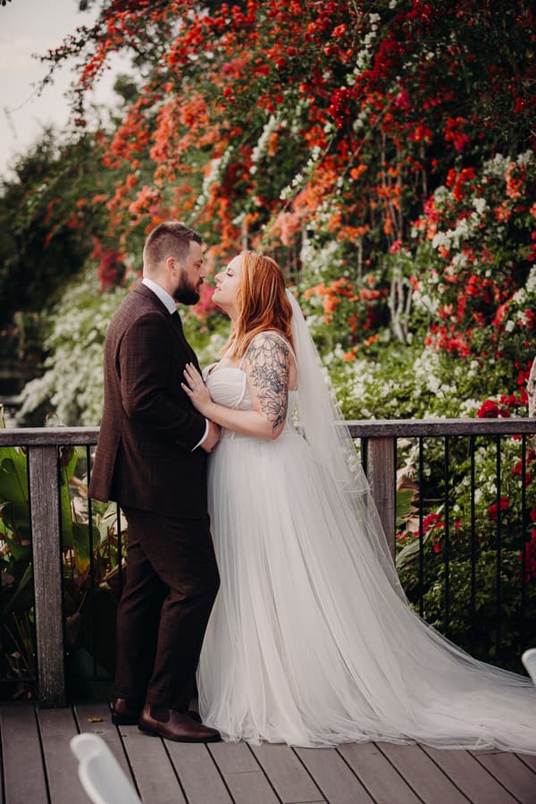 The bride Lilas and groom Kaine stand close together on a wooden deck at Eatons Hill Hotel, surrounded by vibrant red, orange, and white flowers on a lush green backdrop. Lilas wears a strapless white wedding gown with a long veil and visible arm tattoo, while Kaine wears a dark suit and brown shoes.