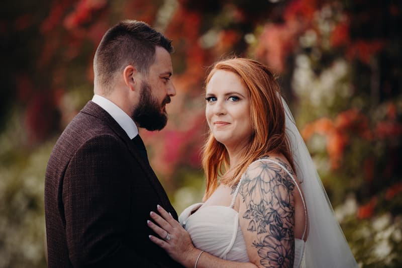 The bride Lilas and the groom Kaine pose together for couple portraits at Eatons Hill Hotel, with Lilas wearing a white wedding dress and veil, showing a floral tattoo on her arm, and Kaine dressed in a dark suit and tie.