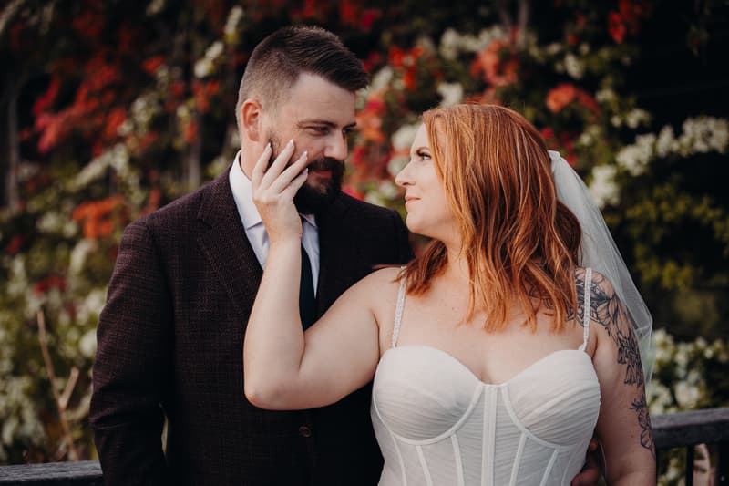 The bride Lilas in a white strapless wedding dress with a veil and visible arm tattoo gently touches the groom Kaine's face as they look at each other in front of a floral backdrop at Eatons Hill Hotel.