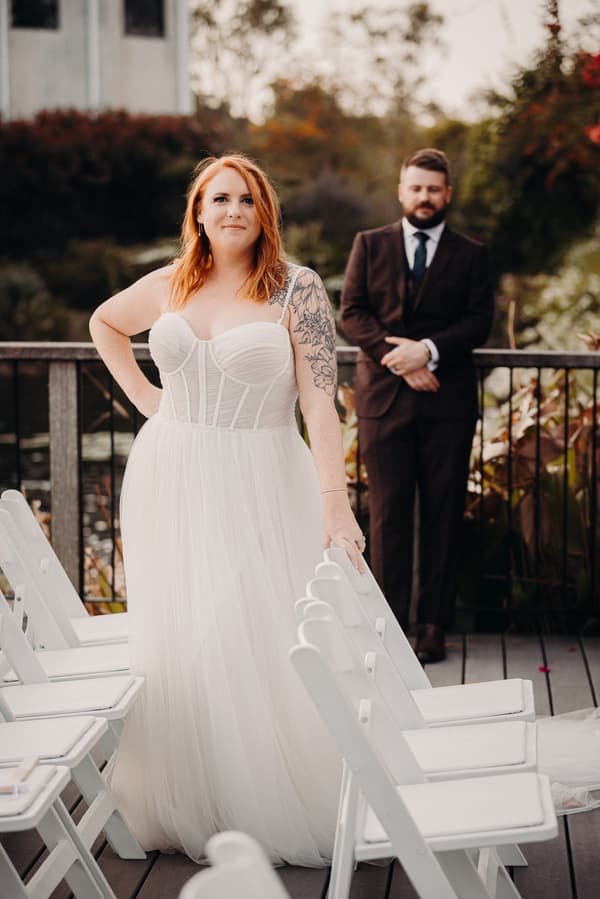The bride Lilas stands in a white wedding gown with a tattooed arm resting on a white chair, while the groom Kaine stands behind her in a dark suit on a wooden deck at Eatons Hill Hotel.