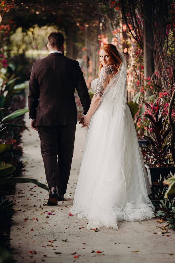 The bride Lilas and groom Kaine walk hand in hand down a garden path at Eatons Hill Hotel. Lilas is wearing a white wedding gown with a veil and has visible tattoos on her arm, looking back towards the camera. Kaine is dressed in a dark suit with his back to the camera.