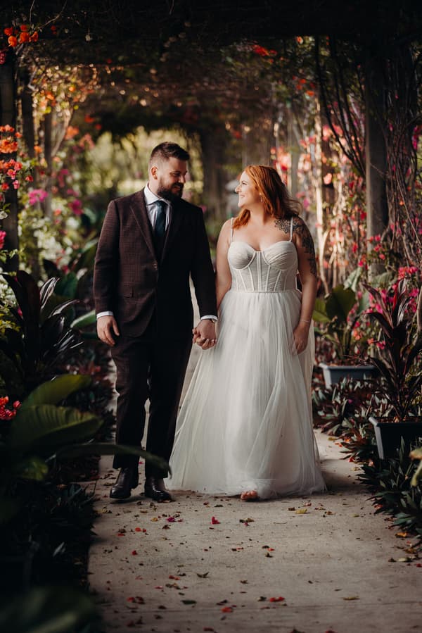 The bride Lilas and groom Kaine walk hand in hand along a flower-lined pathway at Eatons Hill Hotel, surrounded by lush greenery and vibrant flowers.