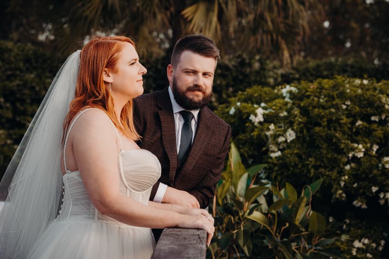 The bride Lilas and the groom Kaine lean on a wooden railing outdoors at Eatons Hill Hotel, surrounded by greenery and white flowers.