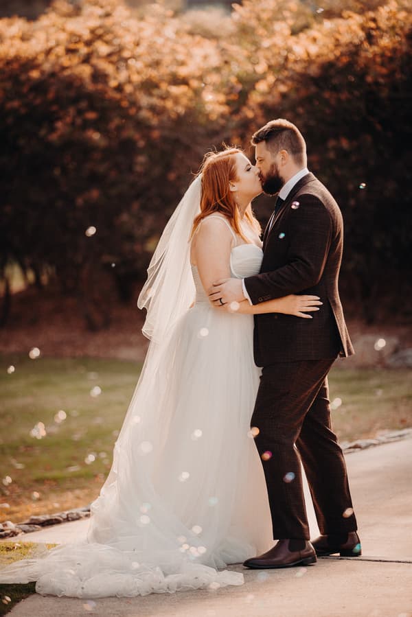 The bride Lilas and groom Kaine share a kiss during their couple portraits session at Eatons Hill Hotel, standing outdoors on a paved path with trees in the background.
